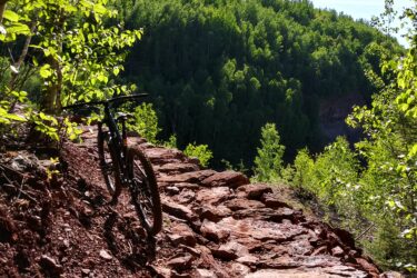 A mountain bike is resting on a rocky trail surrounded by lush green foliage and trees. The scene captures a clear blue sky overhead, with sunlight filtering through the leaves, illuminating the vibrant landscape. The trail appears rugged and adventurous, set against a backdrop of dense forests in the distance. Redhead MTB Park mountain bike trail.