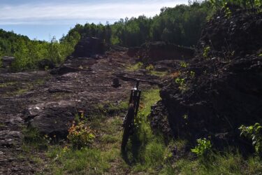 A mountain bike resting on rocky terrain, surrounded by lush greenery and against a bright blue sky with wispy clouds. The sun shines brightly overhead, casting a warm light on the landscape. Redhead MTB Park mountain bike trail.