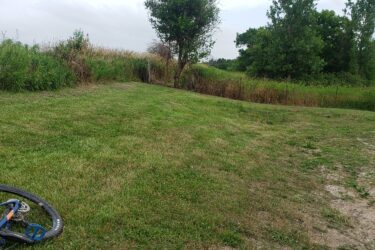 A grassy area with a dirt path leading into a wooded area, accompanied by tall grass and shrubs on either side. In the foreground, a bicycle lies on its side. The sky is overcast, indicating potential rain, and trees can be seen in the background. Lake LaShane Trail mountain bike trail.