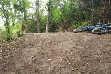 A paused blue mountain bike lies on a dirt trail surrounded by trees and greenery, with a slight incline visible in the background. Lake LaShane Trail mountain bike trail.
