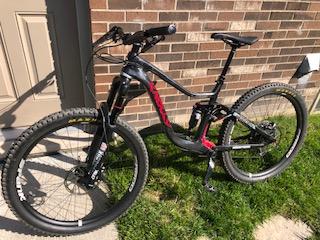 Knolly Warden: A black and red mountain bike parked on green grass in front of a brick wall, with a door visible in the background. The bike features thick tires, a sturdy frame, and a front suspension fork.