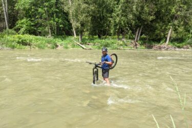 A person in a blue shirt and black shorts is standing in a shallow, muddy river, holding a bicycle. The water reaches mid-calf height, and trees line the riverbank in the background. The scene is sunny and green, suggesting a warm day. Bayfield river mountain bike trail.