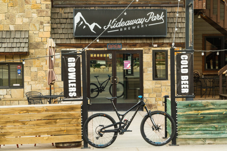 Image of the entrance to Hideaway Park Brewery, featuring a stone building with wooden accents. The brewery sign displays "Hideaway Park Brewery," and there are banners stating "GROWLERS" and "COLD BEER." A black bicycle is parked in front, and outdoor seating is visible on the side. The door is labeled "OPEN."