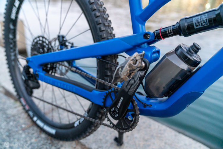 A close-up view of a blue mountain bike's drivetrain, featuring a SRAM crankset, chain, and rear cassette. The bike is equipped with a water bottle mounted on the frame, with a blurred background showing a body of water.