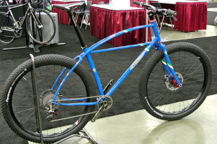 A blue mountain bike with large, thick tires displayed on a stand in a convention hall. The bike features a unique frame design, disc brakes, and a modern gear setup, with a backdrop of tables and other bicycles in a busy exhibit area.