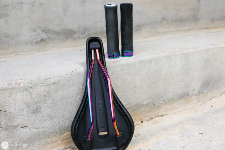 A black bicycle saddle featuring colorful rails sits on a concrete surface, alongside two black bike grips with colorful end caps.