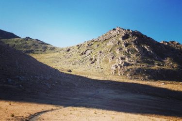 A winding dirt path leading through a grassy valley, flanked by rocky hills under a clear blue sky. The sun casts long shadows across the terrain, highlighting the texture of the ground and the rugged slopes. Stansbury Island Mountain Biking Trail mountain bike trail.