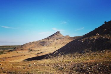 A panoramic view of a rugged landscape featuring a prominent peaked hill, surrounded by rolling hills and expansive plains under a clear blue sky. The foreground showcases a mix of rocky terrain and patches of grass. Stansbury Island Mountain Biking Trail mountain bike trail.