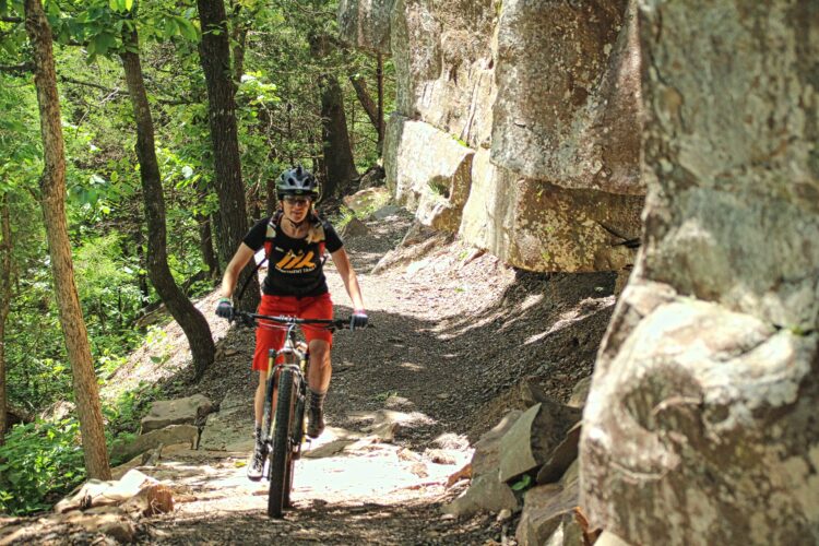 A mountain biker riding on a narrow gravel trail surrounded by lush greenery and rocky outcroppings in a forest setting.