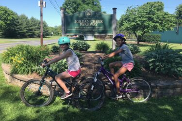 Two children wearing helmets are smiling and posing on their bicycles in front of a sign that reads "Jordan Creek Parkway." The scene captures a sunny day with greenery and flowers surrounding the area, showcasing an inviting park atmosphere. Jordan Creek Parkway mountain bike trail.