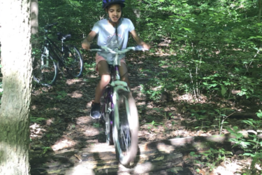A child riding a bicycle along a wooded trail, wearing a helmet and smiling. In the background, another bicycle is parked among trees and greenery, with sunlight filtering through the leaves. Jordan Creek Parkway mountain bike trail.