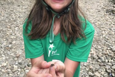 A young girl wearing a turquoise bicycle helmet and a green t-shirt smiles while showing a small creature held in her hands. She stands on a rocky riverbank surrounded by greenery, enjoying a day outdoors. Jordan Creek Parkway mountain bike trail.