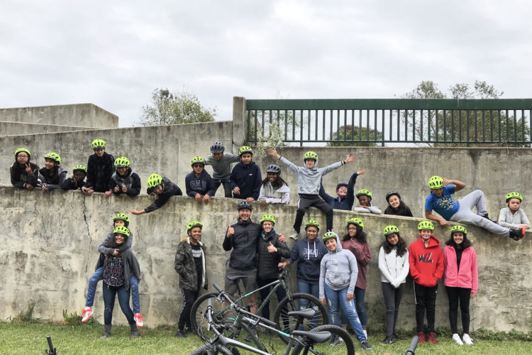 A group of children and adults wearing bright yellow helmets pose cheerfully in front of a weathered concrete wall. Some individuals are leaning against the wall, while others are playfully perched atop it. In the foreground, several bicycles lie on the grass. The scene captures a fun, outdoor biking activity on a cloudy day.