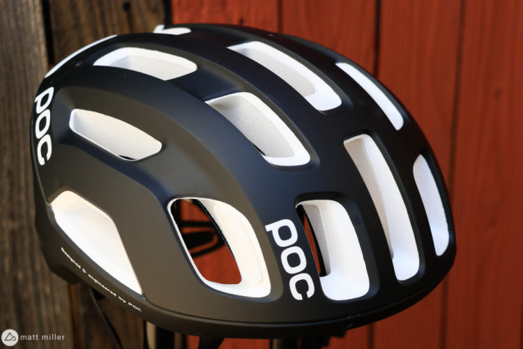 A close-up view of a black and white cycling helmet featuring ventilation slots and the brand name "POC" prominently displayed. The helmet is set against a rustic wooden background.