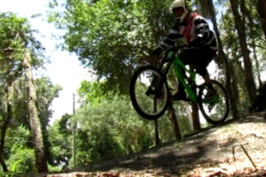 A mountain biker in a helmet and striped jersey is captured mid-air as they jump off a small incline on a wooded trail, surrounded by trees and fallen leaves. Sunlight filters through the foliage, creating a dynamic outdoor scene. Riverbend Nature Park mountain bike trail.