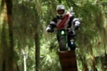 A motocross rider in protective gear is airborne while jumping off a wooden ramp in a forested area, surrounded by trees with hanging moss. The rider is positioned above the ramp, showcasing an action-packed moment. Riverbend Nature Park mountain bike trail.
