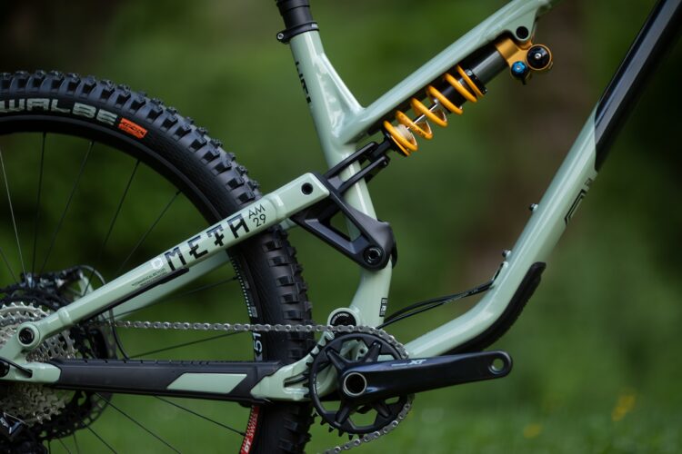Close-up view of a mountain bike's rear suspension system showcasing a green bike frame, yellow shock absorber, and black tire. The image highlights the chain, crankset, and gear system against a blurred green background.