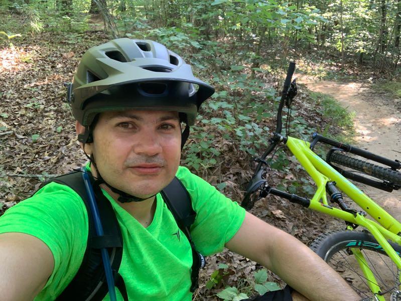 A person wearing a helmet and a bright green shirt sits on the ground next to a mountain bike on a leafy trail surrounded by trees. The scene captures a moment of rest during an outdoor biking adventure. Lakeshore MTB Singletrack mountain bike trail.