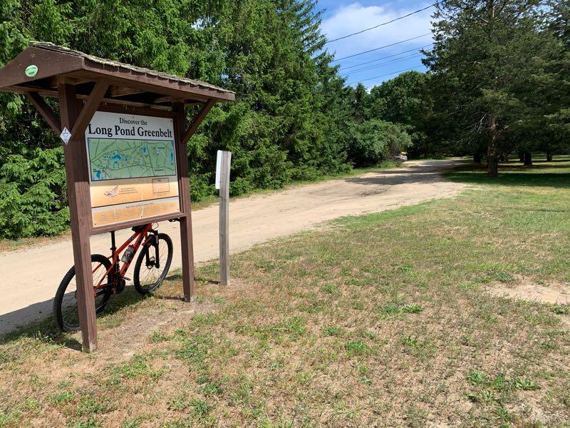 A wooden signpost for the Long Pond Greenbelt is shown next to a dirt path. An orange bicycle leans against the sign, which includes a map and information about the area. Tall trees line the background, and the scene is set on a sunny day with some clouds in the sky. Long Pond Trail mountain bike trail.