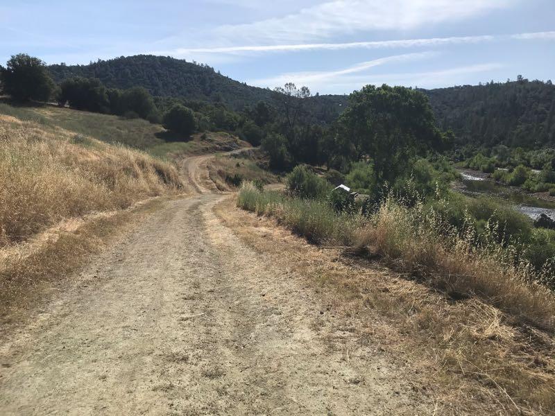 A dirt path winding through a grassy landscape, flanked by bushes and trees, with a distant view of rolling hills and a river below under a blue sky with wispy clouds. Cronan Ranch mountain bike trail.