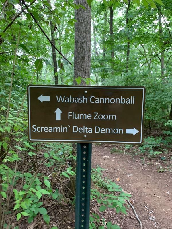 A brown directional sign in a wooded area indicating the paths to "Wabash Cannonball" to the left, "Flume Zoom" straight ahead, and "Screamin' Delta Demon" to the right. Surrounding the sign are green leaves and trees. Percy Warner Mountain Bike Trails mountain bike trail.