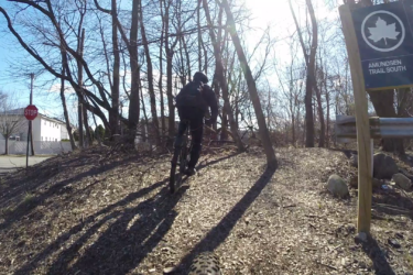 A person riding a mountain bike on a dirt trail surrounded by bare trees, with a stop sign and a trail sign for "Amundsen Trail South" visible in the background. The scene is illuminated by bright sunlight, creating a serene outdoor atmosphere. Richmond Avenue and Forest Hill road mountain bike trail.