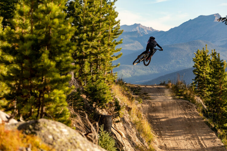 A mountain biker performing a jump on a dirt trail surrounded by pine trees and mountains in the background, with clear blue skies overhead.