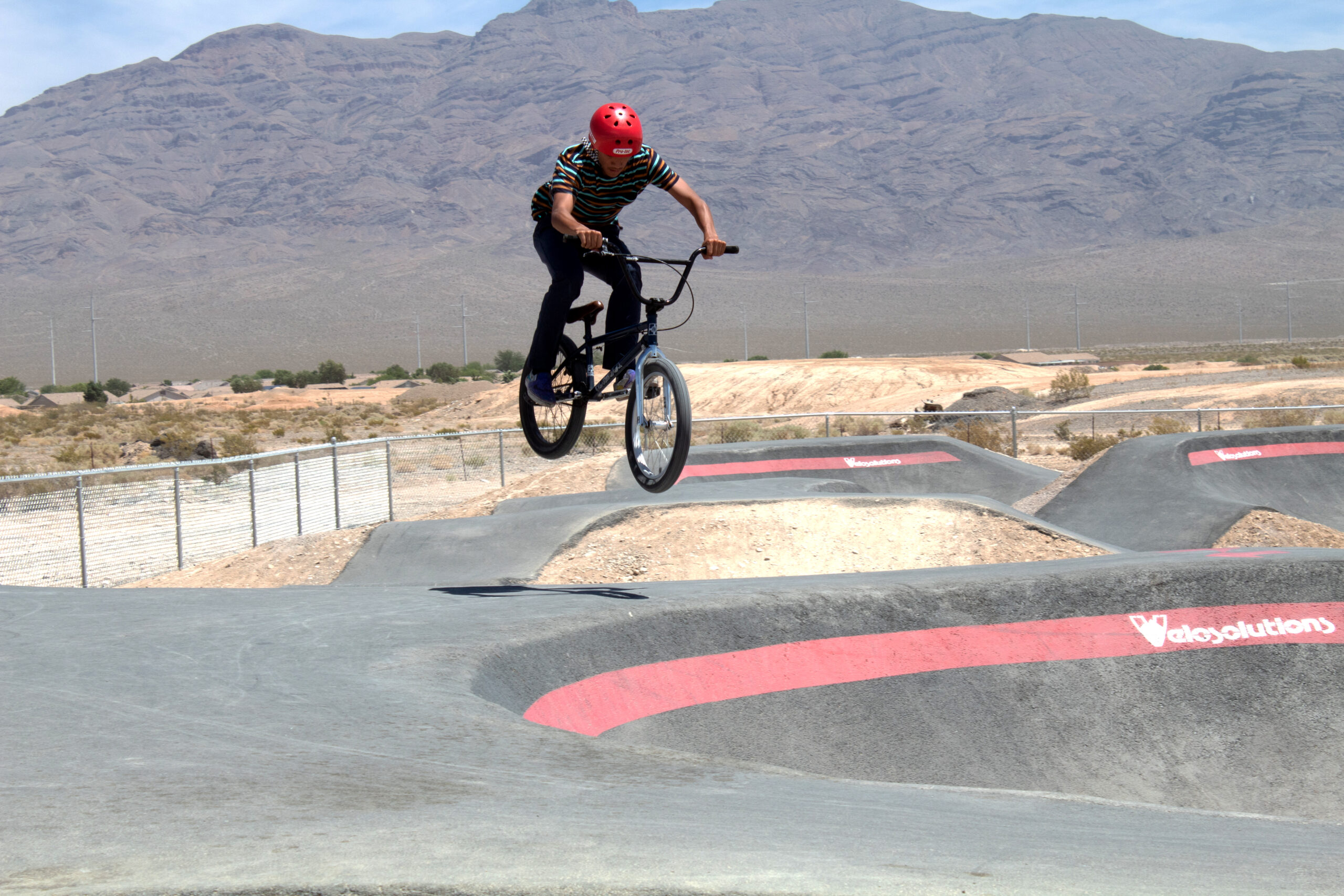 A person wearing a red helmet and a striped shirt is performing a jump on a BMX bike over a dirt track, with a mountainous landscape in the background. The track features a series of bumps and curves, and it is a sunny day. Pump Track mountain bike trail.