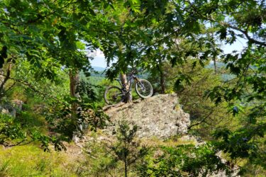 A mountain bike is positioned on a rocky outcrop surrounded by lush green foliage and trees, with distant hills visible in the background under a clear blue sky. Shenandoah River State Park mountain bike trail.