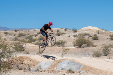 A person wearing a red helmet is mid-air on a BMX bike, performing a jump over a dirt ramp in a desert landscape. The background features dry terrain with sparse vegetation under a clear blue sky. Floyd Lamb Park mountain bike trail.