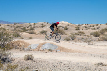 A person wearing a red helmet is performing a jump on a bicycle over a dirt ramp in an arid landscape. The background features sparse vegetation and distant hills under a clear blue sky. Pump Track mountain bike trail.