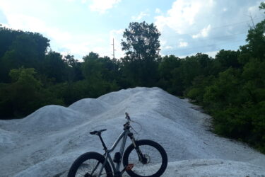 A mountain bike is parked on a white, sandy terrain surrounded by lush green trees under a cloudy sky. The landscape features mounded sand, suggesting an area for off-road cycling or a bike trail. Waggoner's Run Singletrack mountain bike trail.