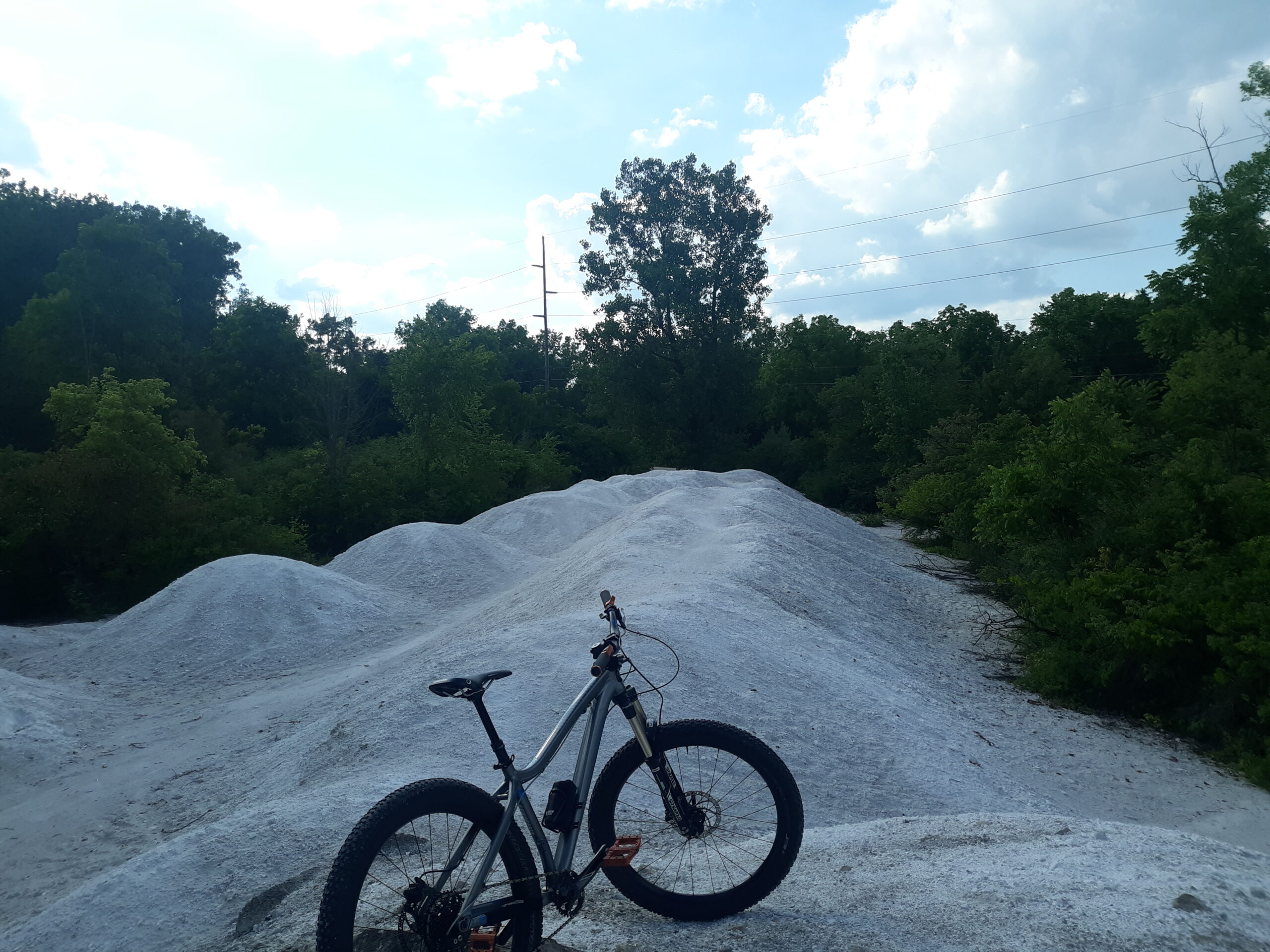 A mountain bike is parked on a white, sandy terrain surrounded by lush green trees under a cloudy sky. The landscape features mounded sand, suggesting an area for off-road cycling or a bike trail. Waggoner