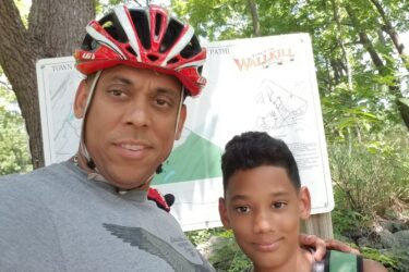 A man wearing a helmet and a gray shirt poses for a selfie with a smiling boy, both standing in front of a map of the Wallkill River area. The background features lush green trees and grass, suggesting an outdoor recreational location. Camp Orange mountain bike trail.
