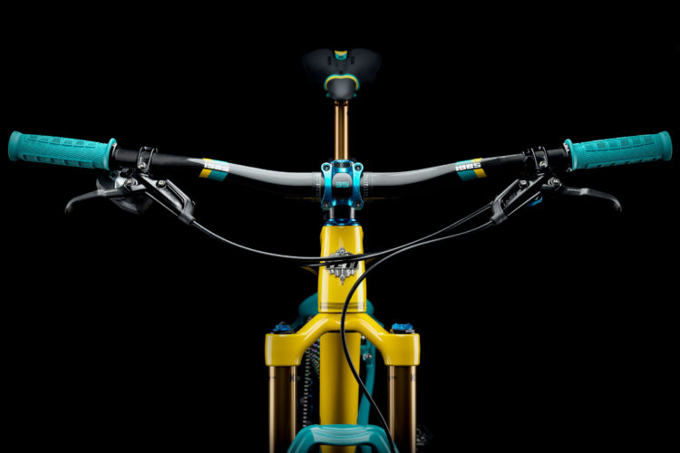 Close-up view of a mountain bike's handlebars and stem against a black background, featuring vibrant teal grips and yellow frame elements, highlighting the intricate design and components.