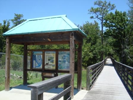 A wooden information kiosk with a blue metal roof, displaying maps and signage, beside a wooden boardwalk leading into a wooded area with trees in the background. Shingle Creek Trail mountain bike trail.