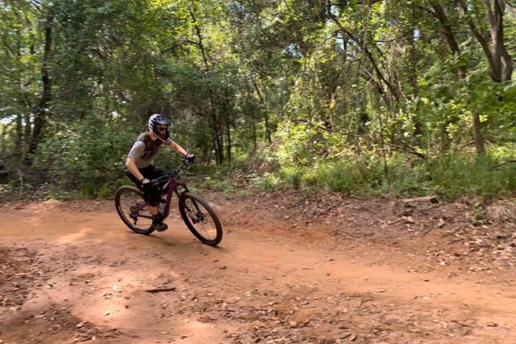 A person riding a mountain bike on a dirt trail through a wooded area, surrounded by greenery, with sunlight filtering through the trees.