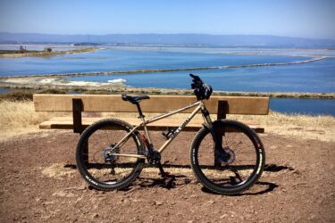 A mountain bike parked next to a wooden bench, overlooking a scenic view of a river and distant hills under a clear blue sky. The foreground features brown soil and grass, while the background showcases the calm water and a winding land formation. Coyote Hills Regional Park mountain bike trail.