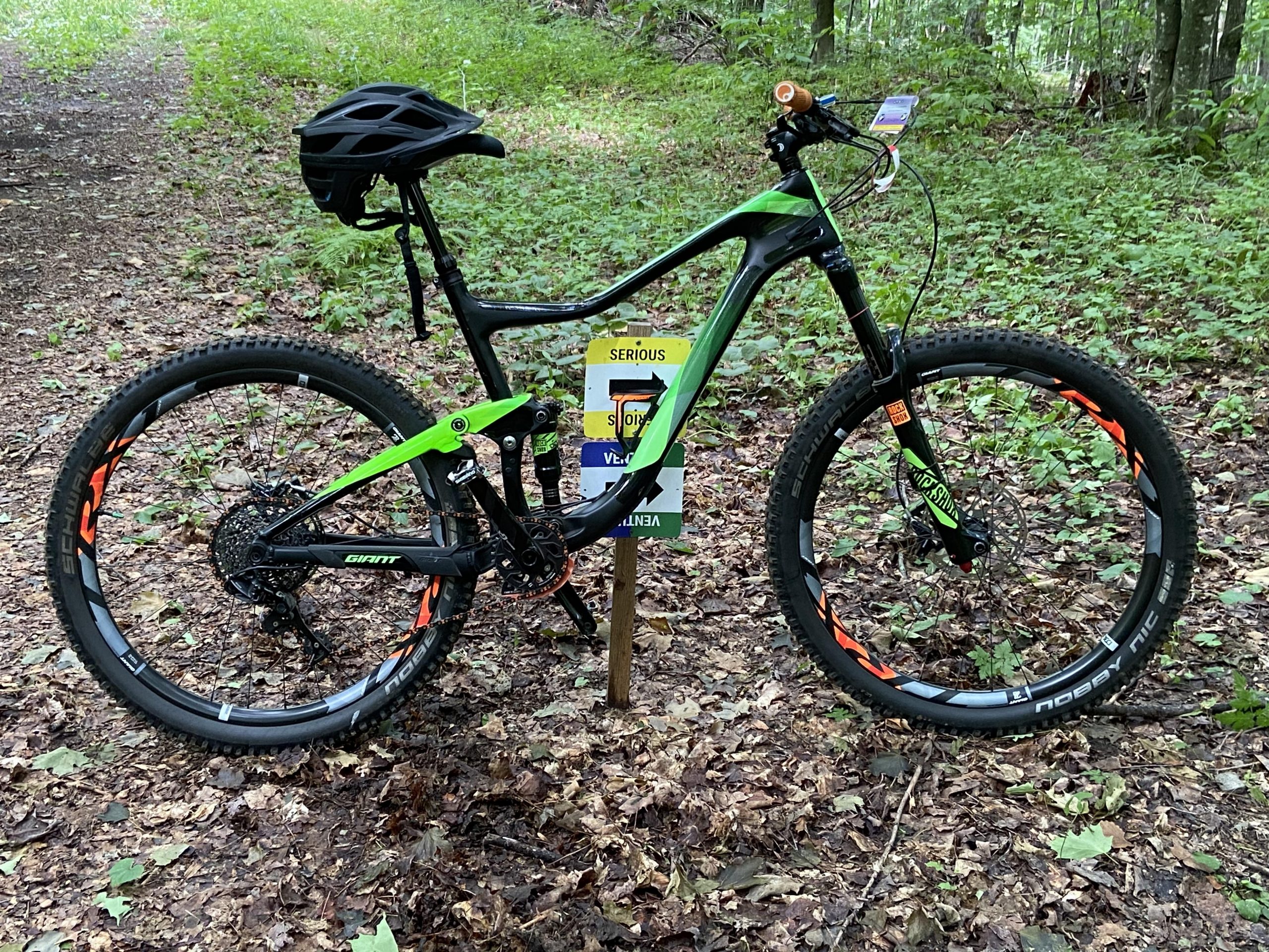 Giant Giant Trance Advanced 27.5 2: A green and black mountain bike leaning against a wooden signpost on a wooded trail. The bike features thick tires and a helmet positioned on the handlebars. The forest floor is covered with leaves and greenery in the background.