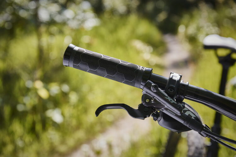 Close-up view of a bicycle handlebar grip featuring a textured, hexagonal pattern. The grip is black, and the bike's brake lever is also visible, with a blurred background of lush greenery suggesting an outdoor setting.