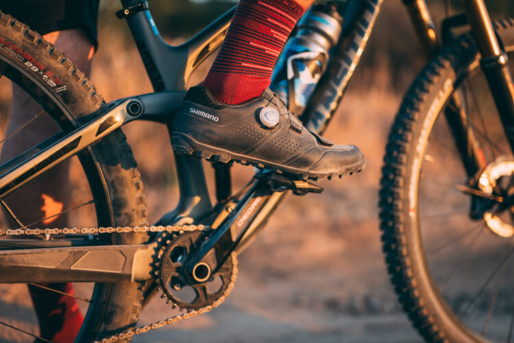 Close-up of a cyclist's foot in a specialized Shimano cycling shoe, positioned on a bicycle pedal. The shoe features a Boa tightening system, and the cyclist is wearing red striped socks. The background shows a blurred mountain bike and a natural outdoor setting in warm lighting.