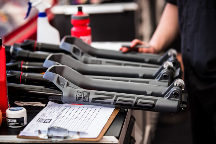 A close-up view of bicycle suspension forks aligned on a workbench, accompanied by maintenance supplies like a spray bottle and a water bottle. A clipboard with handwritten notes is partially visible in the foreground, suggesting ongoing maintenance or inspection work.