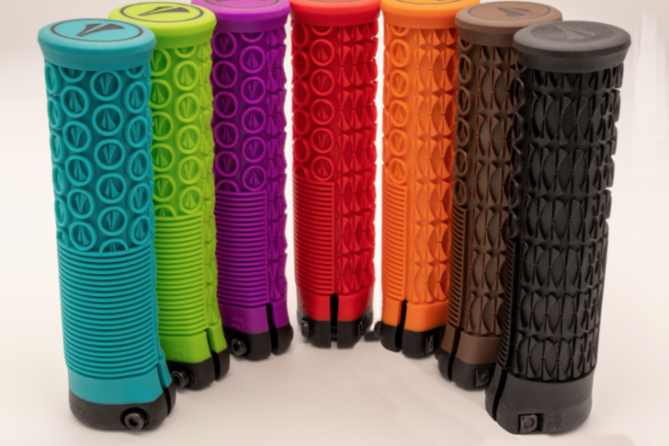 A row of seven colorful bicycle grips arranged vertically, featuring various textures and patterns. The colors include bright teal, green, purple, red, orange, brown, and black. Each grip has a distinct design, showcasing different surface textures for enhanced grip and comfort.