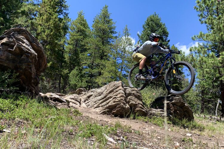 A mountain biker jumps over a large rock formation on a trail surrounded by tall pine trees under a clear blue sky.