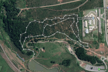 Aerial view of a mapped trail or pathway in a green landscape, showing various distances marked along the route, with nearby roads and buildings visible. The area features grassland and patches of forest, with a river identified to the left and a recreational facility to the right. Trilha do parque da cidade mountain bike trail.