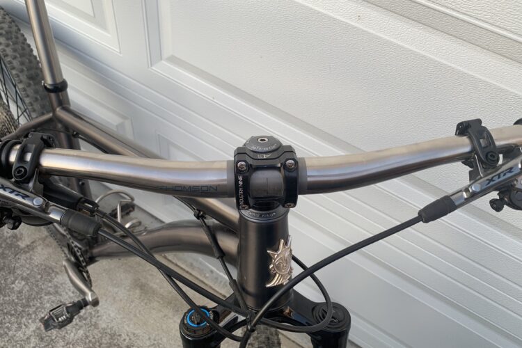 Close-up view of a mountain bike's handlebars and stem, featuring a Thomson stem and XT brake levers. The handlebars are silver and have integrated cables, with an additional decorative element on the bike frame. The background shows a garage door.