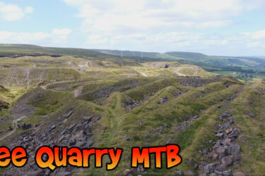 A panoramic view of Lee Quarry, featuring rolling hills and rocky terrain, ideal for mountain biking. The landscape includes a mix of grassy areas and scattered stones, with a clear blue sky and distant hills in the background. A small group of people is visible near the bottom of the image, engaging in outdoor activities. Lee Quarry mountain bike trail.