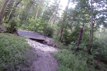 A dirt path winds through a dense forest with lush green trees and underbrush, leading to a small wooden bridge that crosses over a rocky area. The scene is calm and natural, illuminated by gentle sunlight filtering through the tree canopy. Cedarville State Forest mountain bike trail.