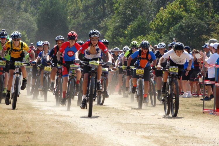 A group of young cyclists in colorful jerseys and helmets races down a dusty trail, with spectators cheering along the sidelines. The atmosphere is energetic, showcasing a competitive mountain biking event. Dust rises as the cyclists pedal forward, creating a dynamic scene filled with enthusiasm and excitement.