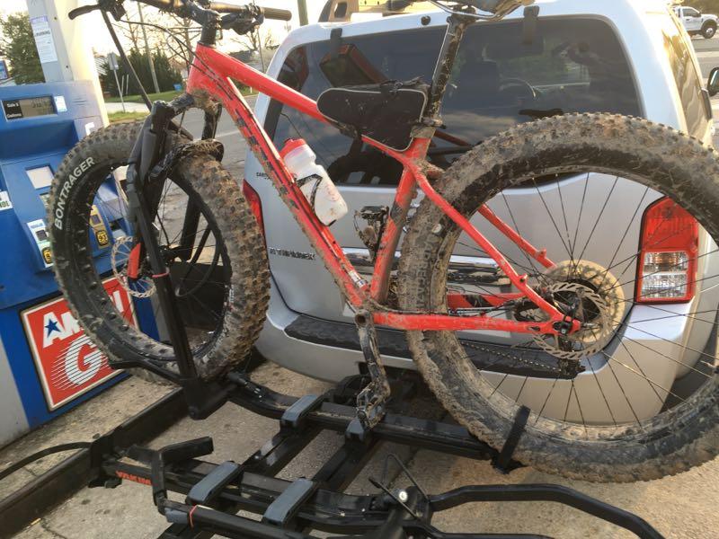 A red mountain bike with muddy tires is secured on a bike rack attached to the back of an SUV. A water bottle is mounted on the bike